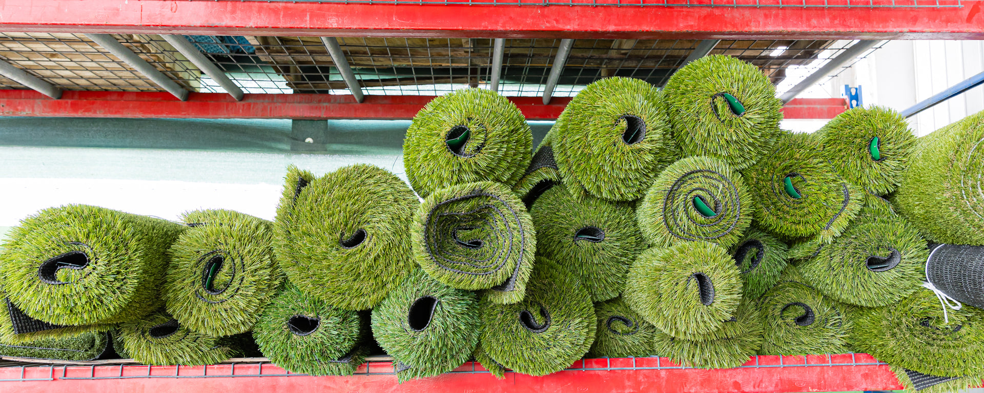 Stack of green artificial grass rolls on a red shelf in a warehouse setting