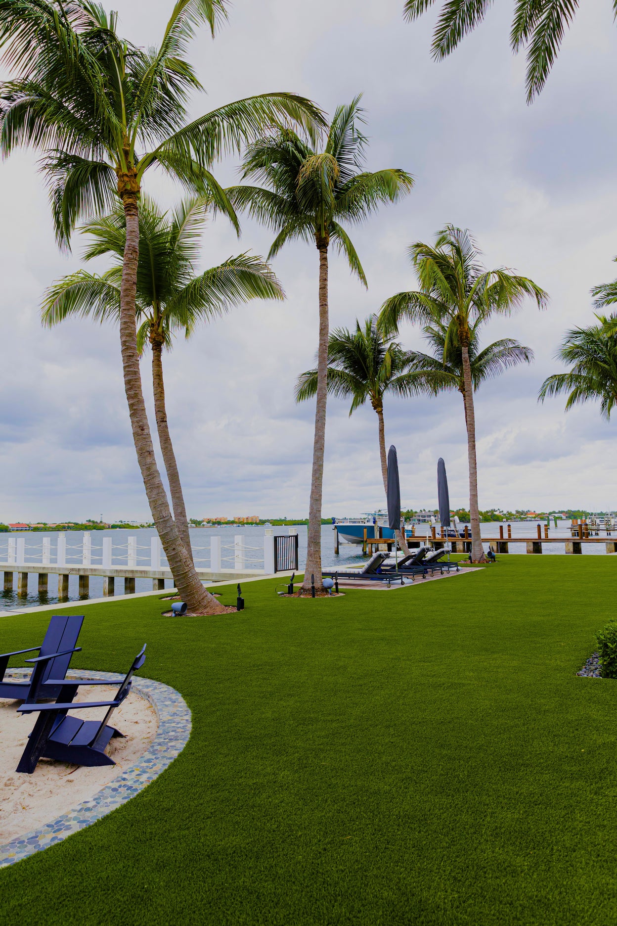 Palm trees and lounge chairs on a grassy area near a waterfront with boats.