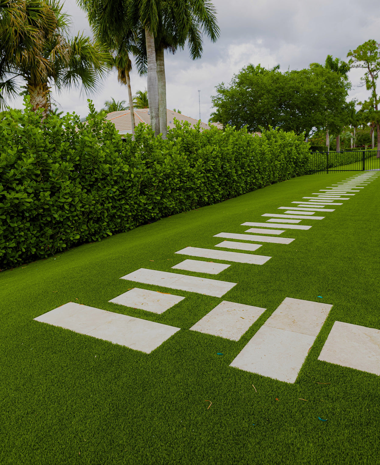 Pathway made of stepping stones on a grassy area with palm trees and hedges in the background.