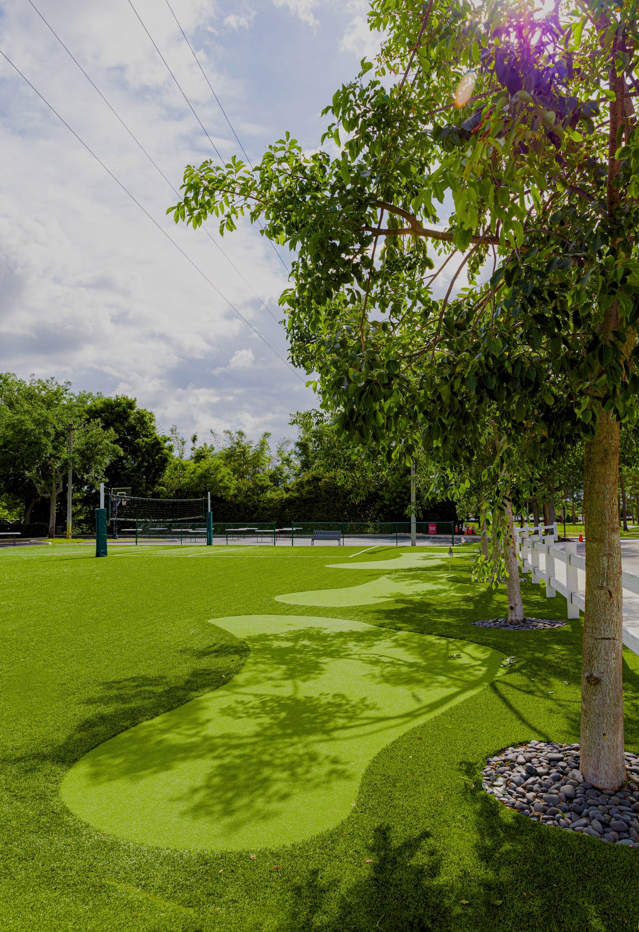 Volleyball court with green grass and trees casting shadows on a partly cloudy day.